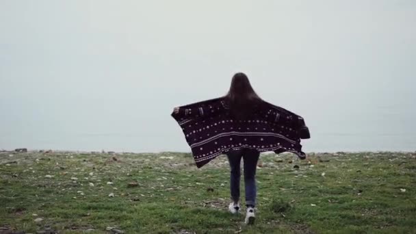 Meisje in een ontwikkelende gebreide poncho loopt rechttrekken haar armen aan de rand van een klif aan zee — Stockvideo