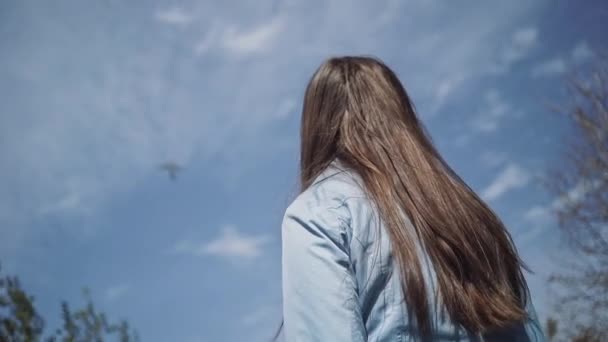 Девушка с длинными светлыми волосами смотрит на самолет, летящий в голубом небе с облаками — стоковое видео
