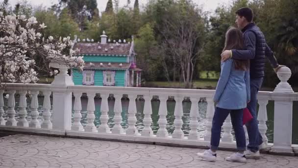 Fagile liten flicka och en lång kille går tillsammans i trädgården med blommande Magnolias längs vallen — Stockvideo