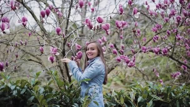 Mooi meisje in een blauwe vacht staat in de buurt van een bloeiende roze kleur magnolia boom en snuiven zijn bloemen, algemeen plan — Stockvideo