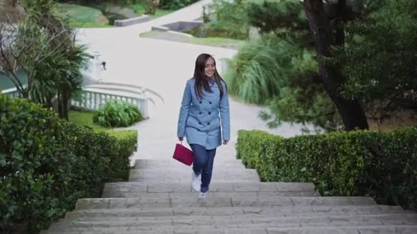 Chica feliz con un abrigo azul y un embrague rosa sube las escaleras de piedra blanca en medio del verde del parque — Vídeo de stock