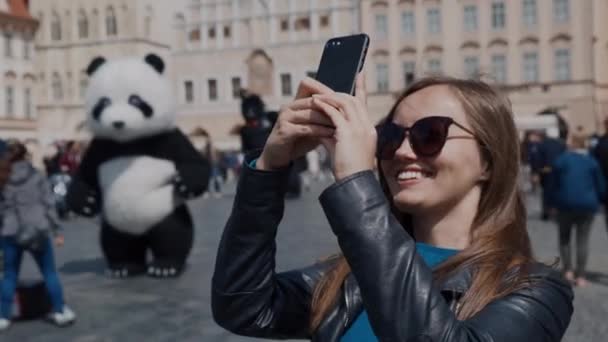 Mooi meisje in zonnebrillen staat op het dorpsplein en foto's van historische gebouwen. Een man in een Panda kostuum op de achtergrond — Stockvideo