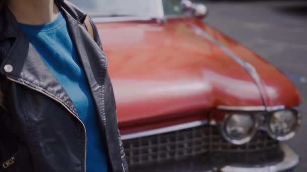 Chica con estilo en gafas de sol y una chaqueta de cuero se encuentra cerca de un coche retro rojo. El cabello se desarrolla maravillosamente en el viento — Vídeo de stock