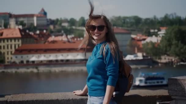 Jeune femme souriante avec des lunettes de soleil debout sur la terrasse promenade de la rivière. Ses cheveux flottent dans le vent fort — Video