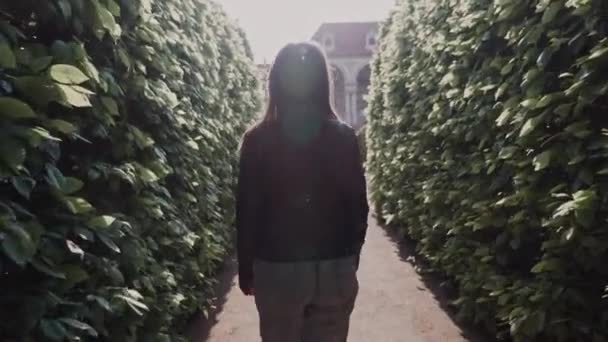 Menina sorridente bonita andando no labirinto do parque na luz do sol da noite. Ela se vira e sorri para a câmera. Visão traseira — Vídeo de Stock