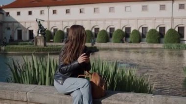 Gölet in yanında güzel bir parkta otururken akıllı telefon kullanarak genç çekici kız. Wallenstein Sarayı ve bulanık arka plan üzerinde balık ve heykeller ile gölet vardır. Güzel peyzaj