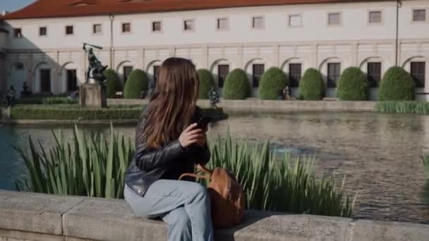 Menina atraente nova usando smartphone enquanto sentado no belo parque à beira da lagoa. Há o Palácio Wallenstein e a lagoa com peixes e esculturas sobre fundo azul. Belo paisagismo — Vídeo de Stock