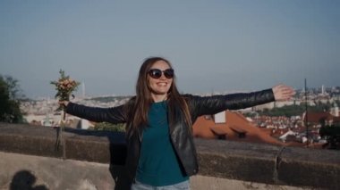 güneş gözlüğü ve deri ceket gülümseyen kız güneşli bir günde uzanmış kolları ile evlerin çatıları düzeyinde taş parapet yanında duruyor. Küçük buket kızlar elinde