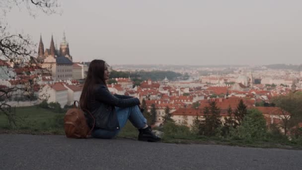 Schönes Mädchen mit Rucksack in Jeans und Stiefeln sitzt nachdenklich auf einer Aussichtsplattform mit Blick auf Prag. Mädchen links im Rahmen — Stockvideo