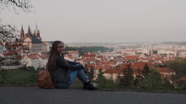 Schöne glückliche Mädchen mit Rucksack sitzt auf der Strecke auf dem Berg mit malerischem Blick auf das alte Prag — Stockvideo