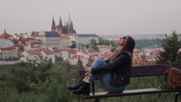 Красивая девушка в солнечных очках, кожаной куртке и сапогах сидит ногами на скамейке с видом на традиционные чешские крыши под вечерним солнцем — стоковое видео