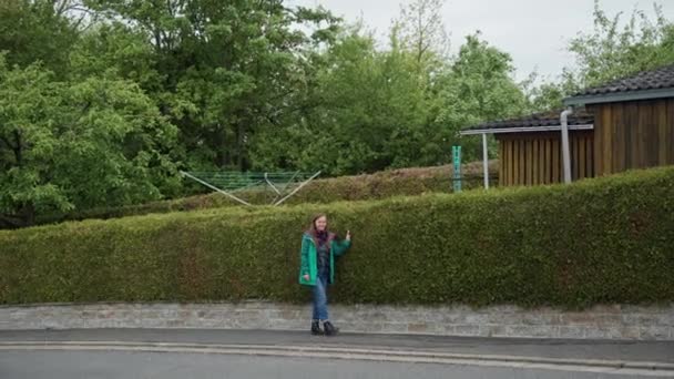 Almanya banliyösünde yeşil çit boyunca yürüyen yeşil ceket, kot pantolon ve çizmesevimli kız — Stok video
