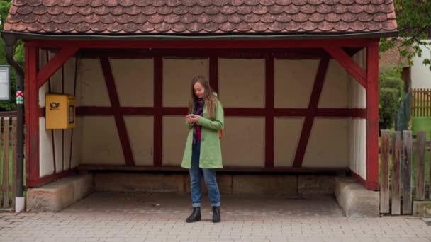 Piękna kobieta w zielonym płaszczu i plecaku przy użyciu smartfona czekając na autobus w pobliżu przystanku autobusowego w stylu fachwerk, Bawaria, Niemcy — Wideo stockowe