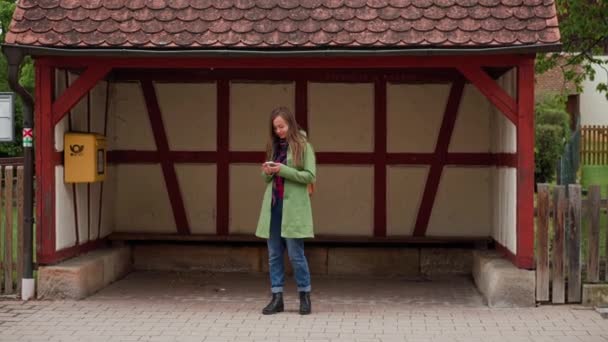 Schöne Frau in grünem Mantel und Rucksack mit Smartphone beim Warten auf einen Bus in der Nähe einer Bushaltestelle, Bayern, Deutschland — Stockvideo