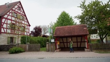 Yeşil ceket ve sırt çantası yalnız kadın Fachwerk tarzı otobüs durağı, Bavyera, Almanya, genel plan yakınında bir otobüs bekliyor