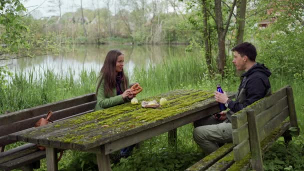 女の子と男は、サーモスでリンゴ、ベンゼルとホットティーとピックニックを持っています。彼らは緑の苔で覆われた古い木製のベンチとテーブルに座っている — ストック動画