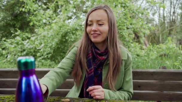 Красивая девушка сидит на деревянной скамейке для пикника, берет бризель и смотрит через нее в камеру и улыбается — стоковое видео