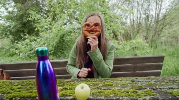 女孩坐在木菜豆青苔覆盖，并通过布雷泽尔看着相机和微笑。紫色热水瓶和绿色苹果在前景 — 图库视频影像