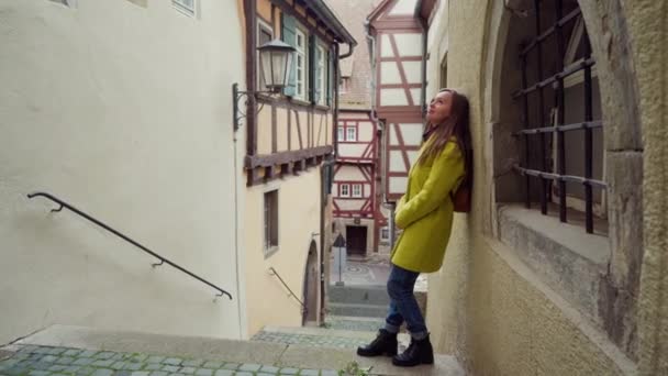 Mladá žena ve žlutém kabátě stojí na stěně domu v malém německém městě. Šťastná dívka, která se baví chodit v roztomilým malém, hrázděném městě se starými domy ve Schwabisch Hall v Německu — Stock video