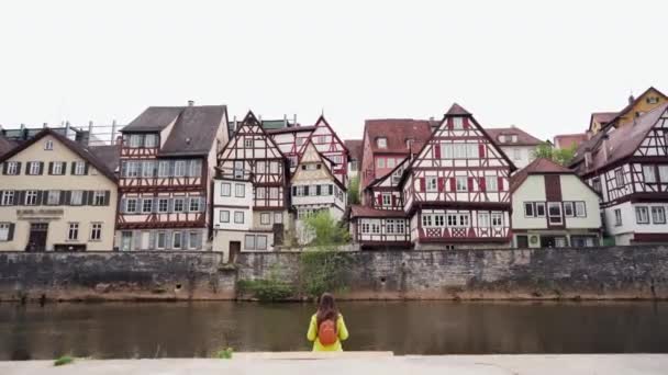 Langhaarige Frau in gelbem Mantel mit Rucksack sitzt mit dem Rücken zum Wasser vor dem Hintergrund traditioneller Fachwerkhäuser. Schwäbisch Hall. Deutschland. Allgemeiner Plan — Stockvideo