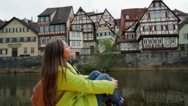 Bella donna in cappotto giallo con zaino siede vicino all'acqua. Ragazza chiuso gli occhi in piacere. Tradizionali case a graticcio della Germania con riflessi sul canale sono sullo sfondo. Tempo di relax — Video Stock