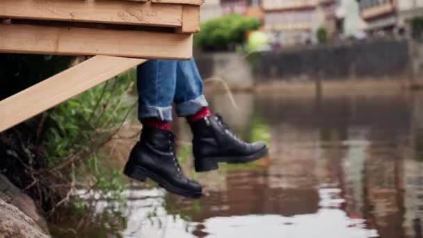 Dames benen in jeans en laarzen hangen over de rand van een houten pier — Stockvideo