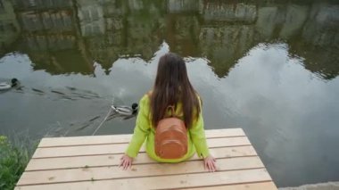 Sırt çantası ile sarı ceketli uzun saçlı kadın nehir veya gölet ahşap iskelede oturur ve ördekler yüzmek görünüyor. Geleneksel Alman yarı ahşap evleri suya yansır. Geri görünüm