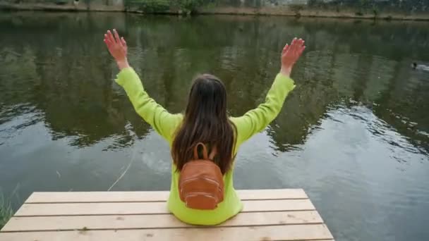 Langhaarige Frau in gelber Jacke mit Rucksack sitzt auf einem hölzernen Steg am Fluss oder Teich und winkt. Die traditionellen deutschen Fachwerkhäuser spiegeln sich im Wasser. zurück — Stockvideo