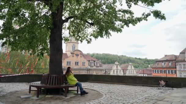 Menina bonita em casaco amarelo senta-se sozinho em um banco no parque ou jardim sob uma árvore grande alta com vista para edifícios coloridos da Baviera — Vídeo de Stock