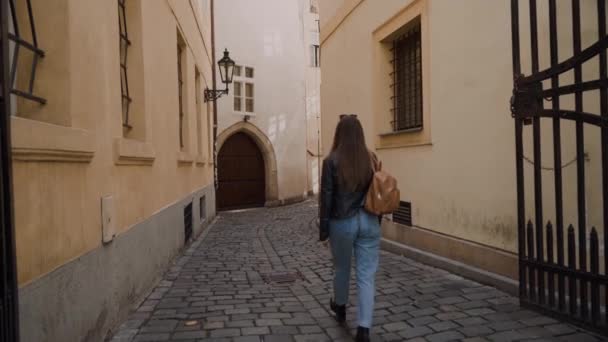 Девушка в джинсах и с рюкзаком идет по красивой узкой старой улице. На стене установлена уличная лампа, арка, которая видна издалека. Вид сзади — стоковое видео