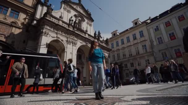 ПРАГА, 15 МАЯ 2019: девушка стоит неподвижно на оживленной площади в Праге, вокруг много людей идет о своем бизнесе, трамваи и автомобили проходят — стоковое видео