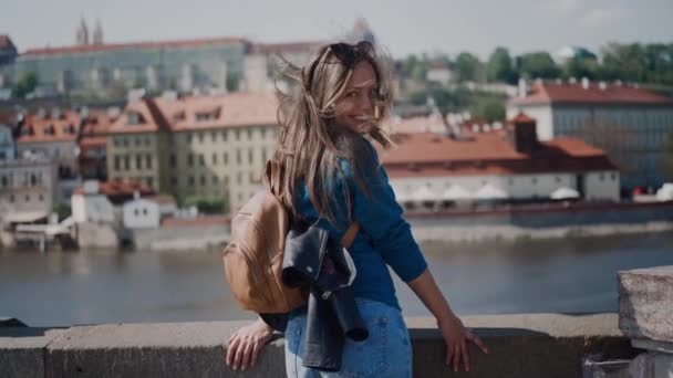 Menina com mochila fica na ponte, sorri e olha para a câmera, no fundo do rio e vista pitoresca da cidade velha. O vento infla muito o cabelo das meninas — Vídeo de Stock