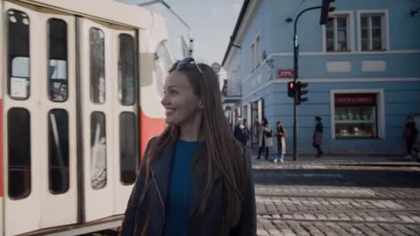 Beautiful girl stands at intersection on street in the background of passing red retro tram and smiles, Prague, Czech Republic — Stock Video