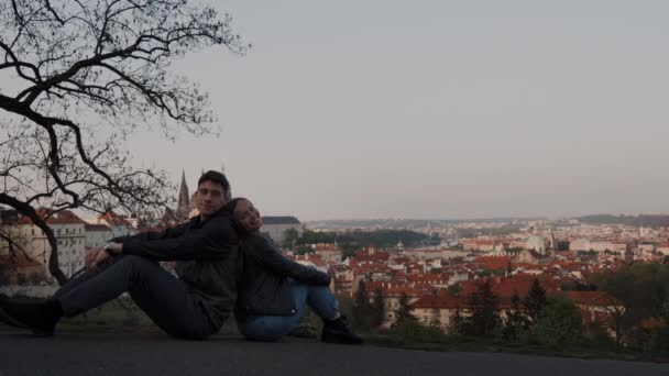 Casal amoroso sentar de volta para trás no deck de observação com vista panorâmica de Praga, eles alegremente olhar para a câmera e mostrar gostos — Vídeo de Stock