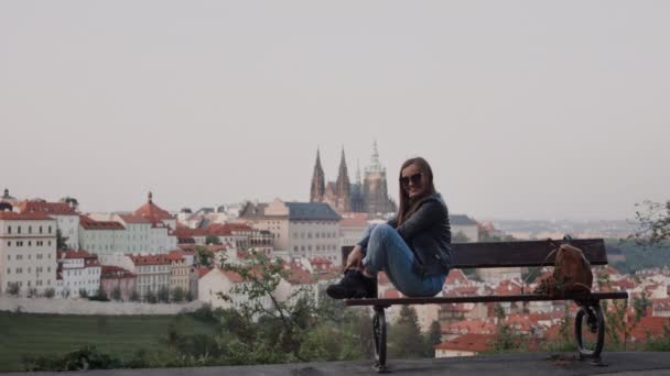 Fröhliches Mädchen mit Sonnenbrille sitzt mit Beinen auf Bank. es gibt traditionelle tschechische Dächer im Hintergrund — Stockvideo