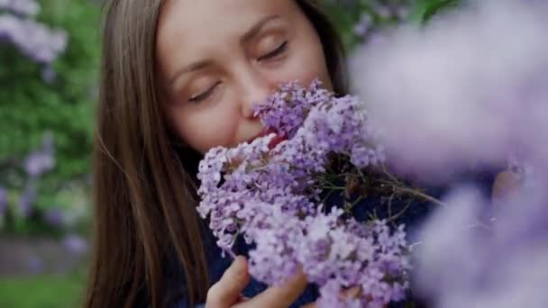 Chica en acogedor suéter olfatea exuberante rama de lila y ella sonríe, en primer plano — Vídeo de stock
