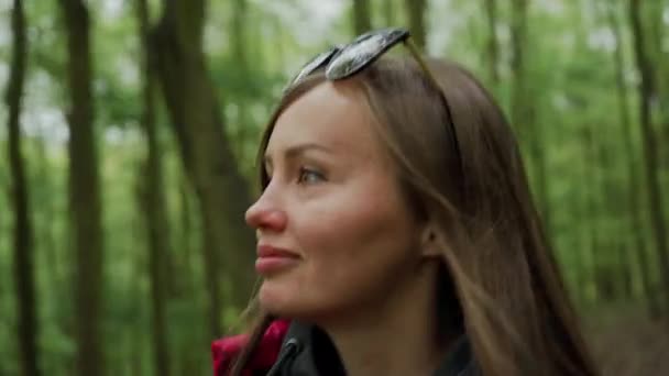 Mulher muito sorridente em jaqueta vermelha com mochila caminha na floresta densa, olha em volta, gosta da beleza da natureza. Caminhadas, hobby, relaxamento, tempo de recreação — Vídeo de Stock