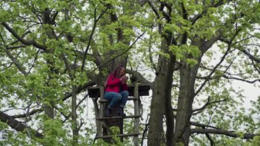 Kırmızı ceketli gülümseyen kadın güçlü ağaç yaprakları ahşap bir merdiven üstüne oturur ve bir akıllı telefon kullanır