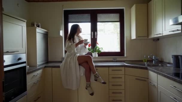 Muchacha sonriente atractiva en albornoz blanco y calcetines de lana bebe café o té y sentado en la mesa de piedra en la acogedora cocina por la mañana. Hermosa cocina blanca y una ventana con una flor en — Vídeo de stock