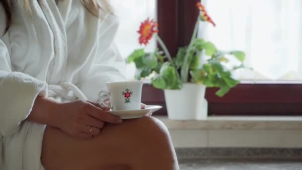 Nahaufnahme: Frau im weißen Bademantel sitzt am Fenster und hält eine weiße Tasse Tee oder Kaffee an ihren Beinen. Kaffeepause — Stockvideo