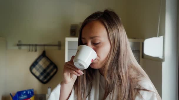 Hermosa chica de albornoz blanco bebe café o té y se sienta en la mesa de piedra en la acogedora cocina por la mañana. Retrato — Vídeos de Stock