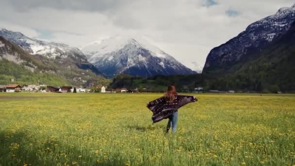 Bakifrån. Lycklig kvinna i mönstrad kappa körs med utsträckta armar över maskrosor fält i natursköna Snow Mountain Valley. Flickan snurrar med glädje. Allmän plan — Stockvideo