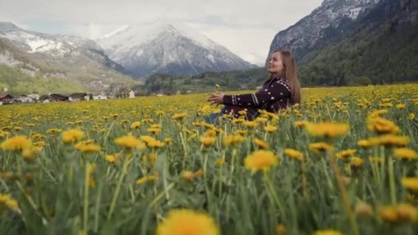 Eine fröhliche junge Frau sitzt auf der Frühlingslöwenzahnwiese, majestätische Berge sind im Hintergrund zu sehen. Seitenansicht — Stockvideo