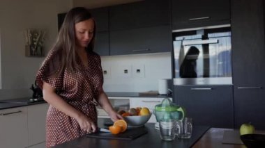 Kahverengi sabahlık gülümseyen kadın taze meyve suyu yapmak için mutfakta siyah tahta üzerinde bıçak ile portakal keser. Genel plan