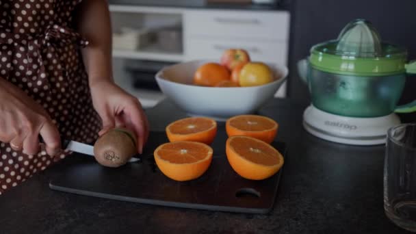 Las manos de una mujer o cocinar kiwi cortado por cuchillo en la tabla de cortar negro con naranjas picadas. De cerca. — Vídeo de stock