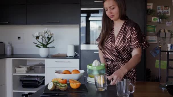 Giovane donna che versa succo d'arancia appena fatto da uno spremiagrumi in un bicchiere in cucina — Video Stock