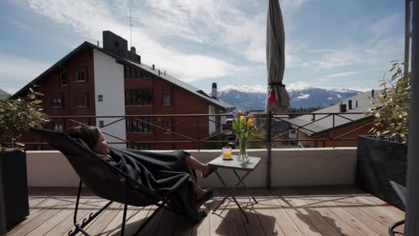 Jovem bela mulher se saciando ao sol deitado em uma cadeira em uma xadrez no terraço com vista para a montanha — Vídeo de Stock