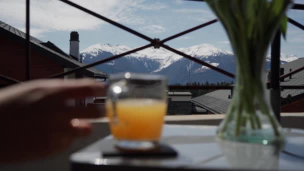 Mano femminile che prende il vetro con succo d'arancia fresco dal tavolo sullo sfondo pittoresco della montagna — Video Stock