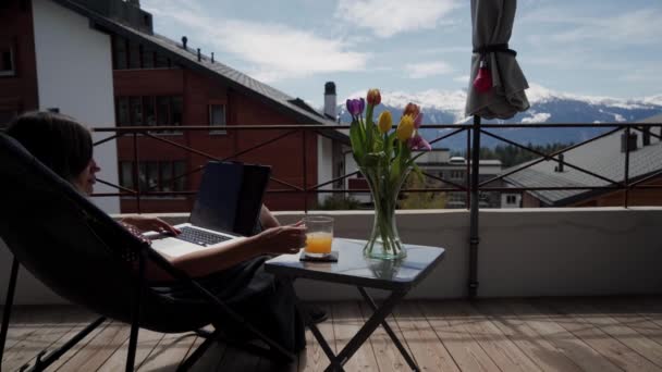 Mujer joven que trabaja fuera en el ordenador portátil y beber jugo de naranja en la terraza con hermosa vista a la montaña. Ramo de tulipanes coloridos está junto a la mesa — Vídeos de Stock