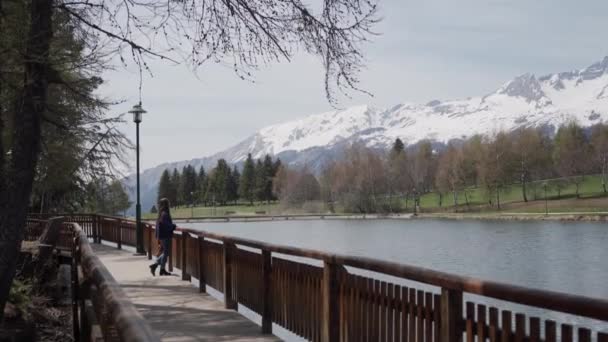 Gyönyörű lány áll a fa sétány terasz és élvezi a festői kilátás nyílik a hegyi tó és a svájci Alpokban. Általános terv — Stock videók